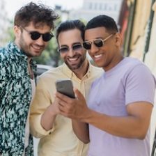 Positive interracial friends in sunglasses using mobile phone on urban street in summer