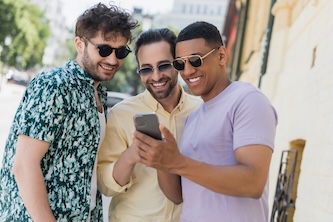Positive interracial friends in sunglasses using mobile phone on urban street in summer